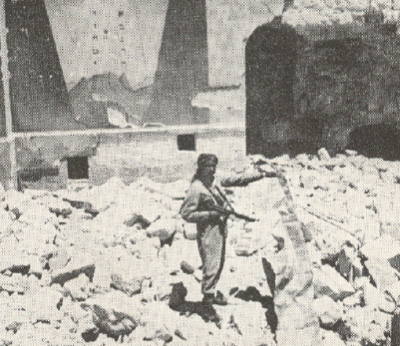 חייל הלגיון הערבי עומד בתוך הריסות בית הכנסת החורבה ומחזיק בידו מגילה של ספר תורה